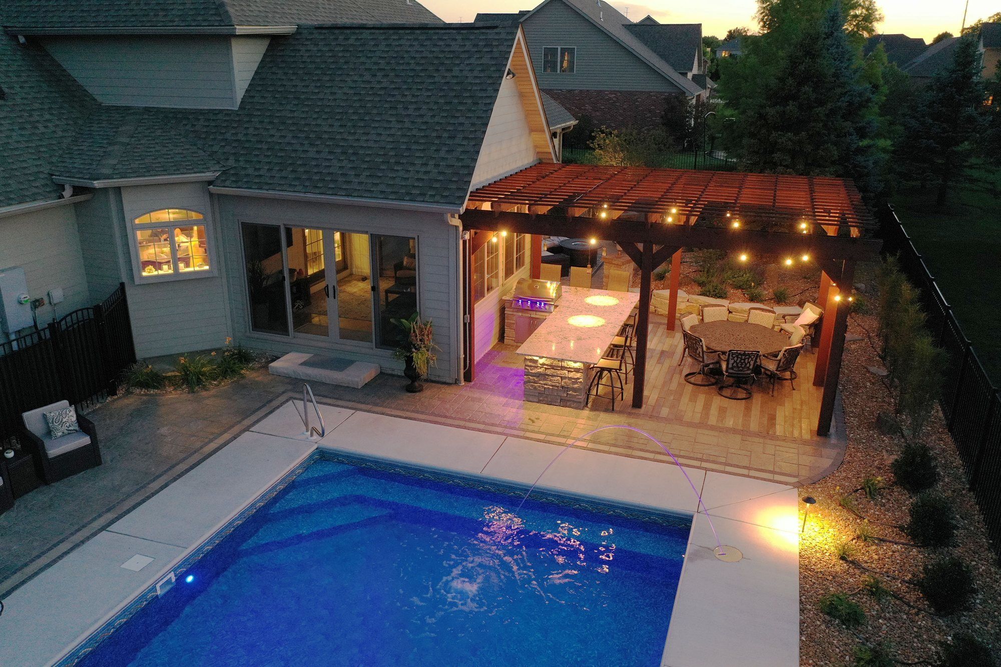 Aerial view of a backyard pool, paver deck and lit pergola at dusk
