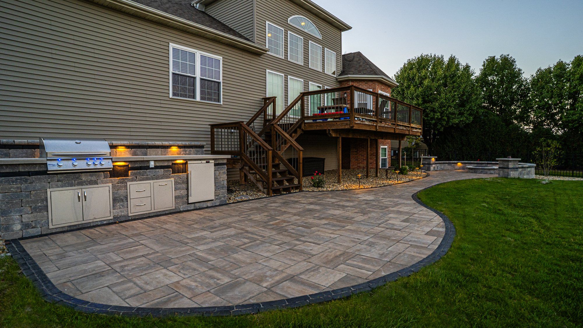 Curved backyard paver patio with a built-in outdoor kitchen below a deck at a Bloomington home