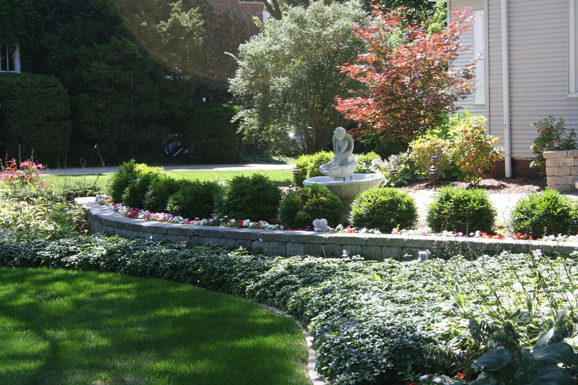 Curved block seat wall with a fountain, boxwoods and flower beds in front of a home