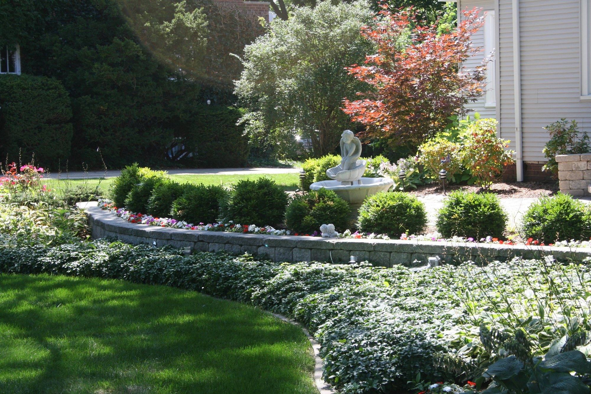 Curved block seat wall with a fountain, boxwoods and flower beds in front of a home