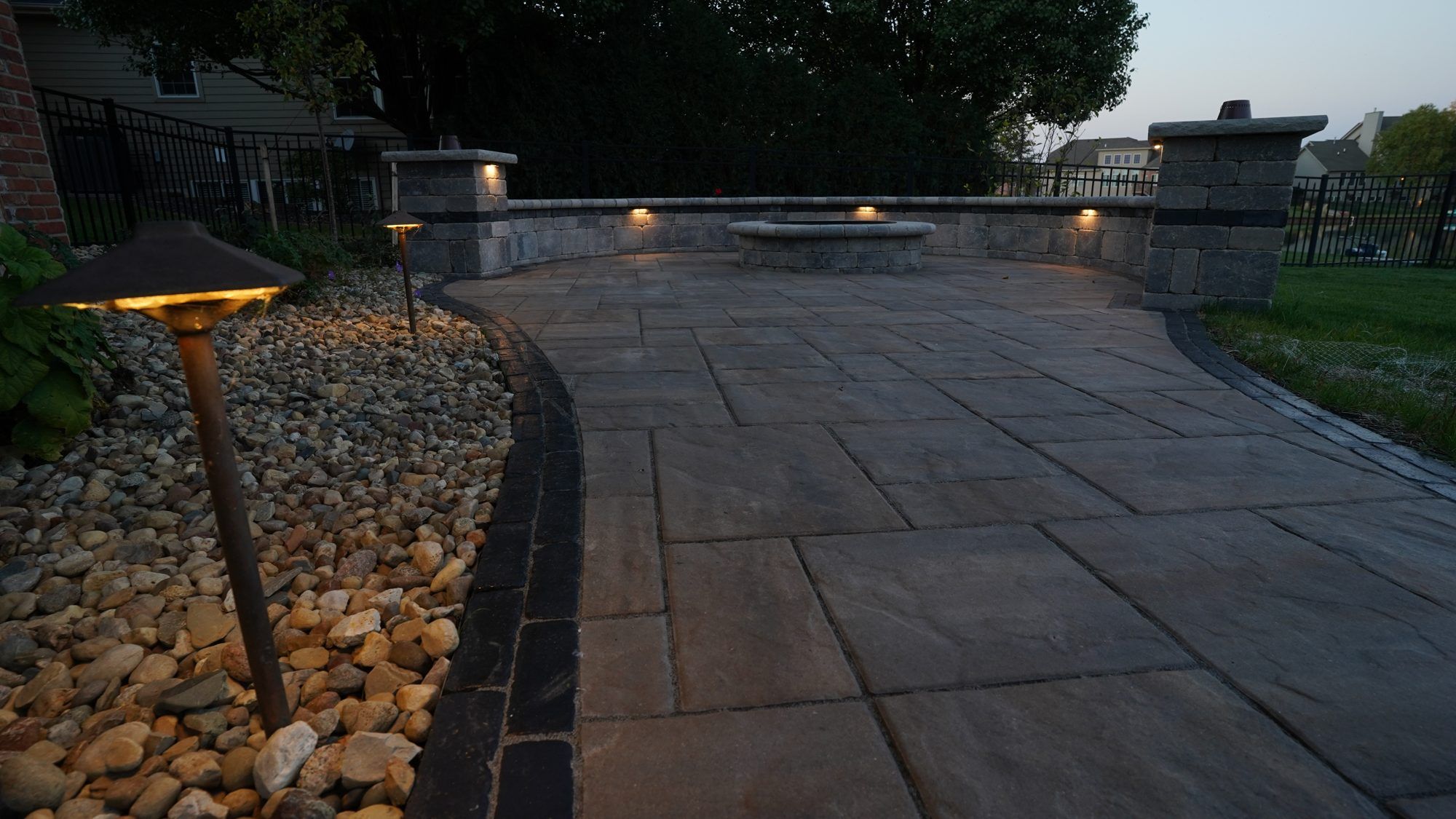 Path light in a river-rock border beside a paver patio and seat wall at dusk