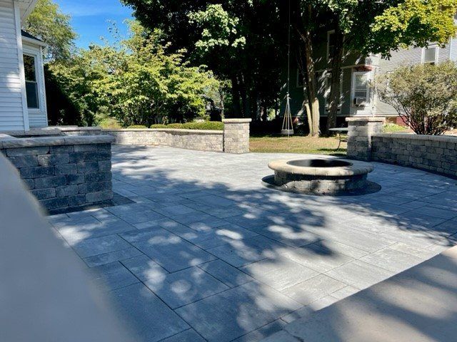 Paver patio with a fire pit, seat wall and stone pillar in a shaded backyard