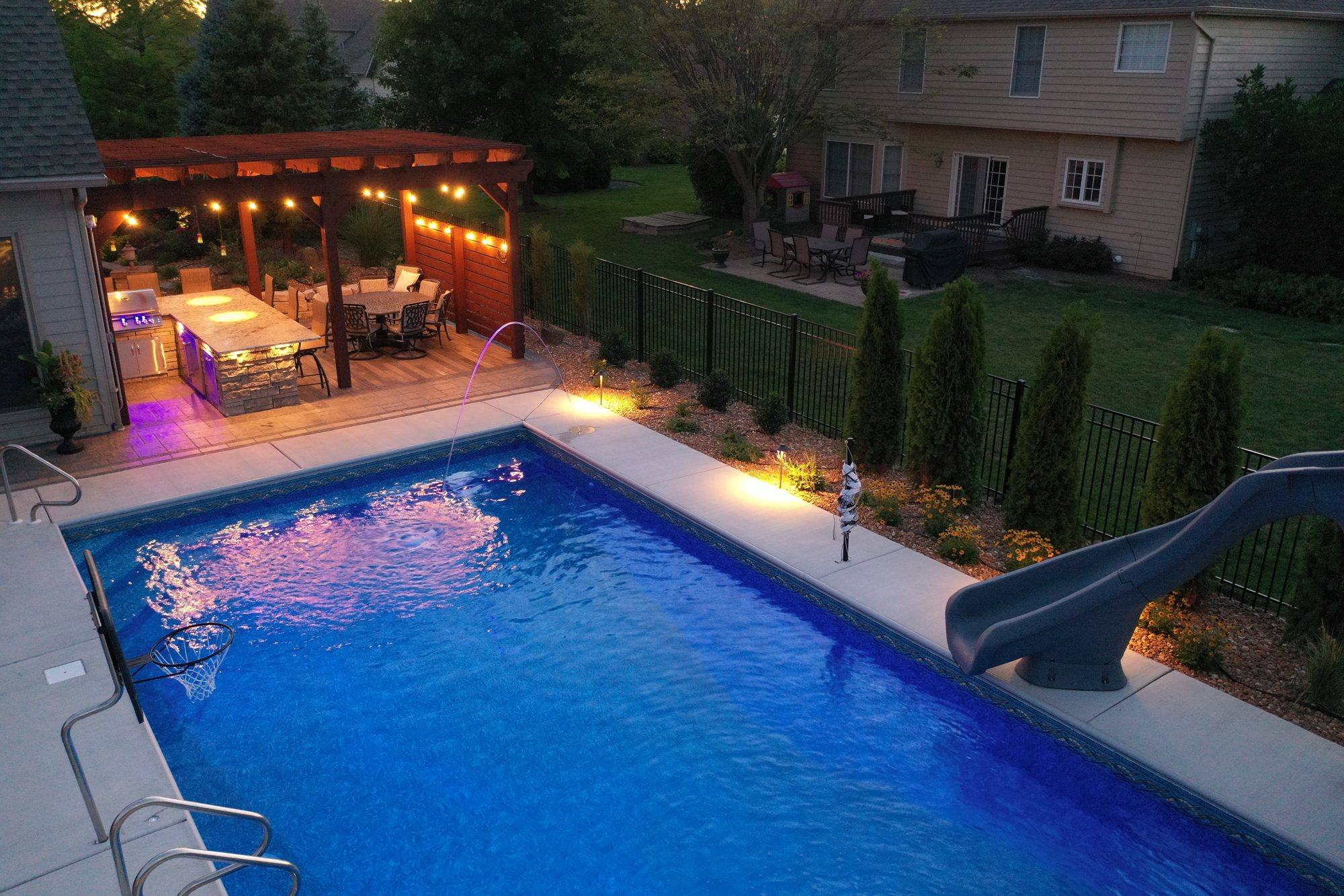 Pool with paver deck, landscape lighting and a lit pergola at dusk