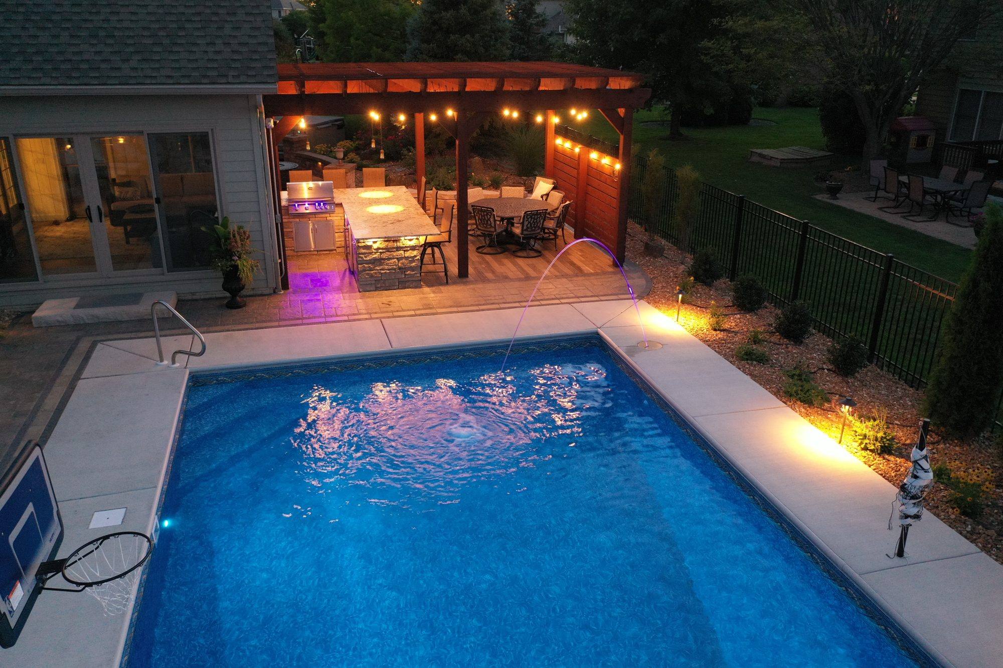 Pool, paver deck and a pergola-covered outdoor kitchen lit up in the evening
