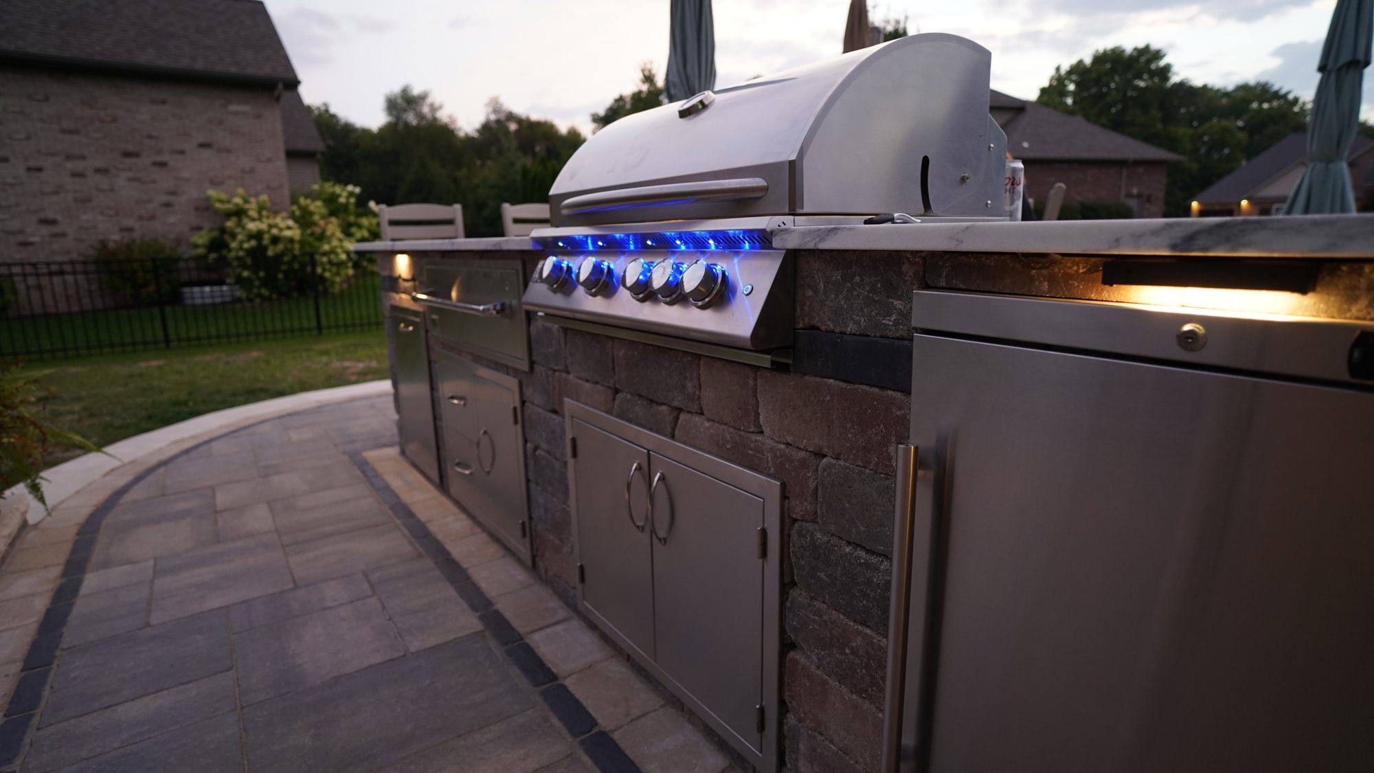 Stainless built-in grill and cabinets in a stone outdoor kitchen at dusk