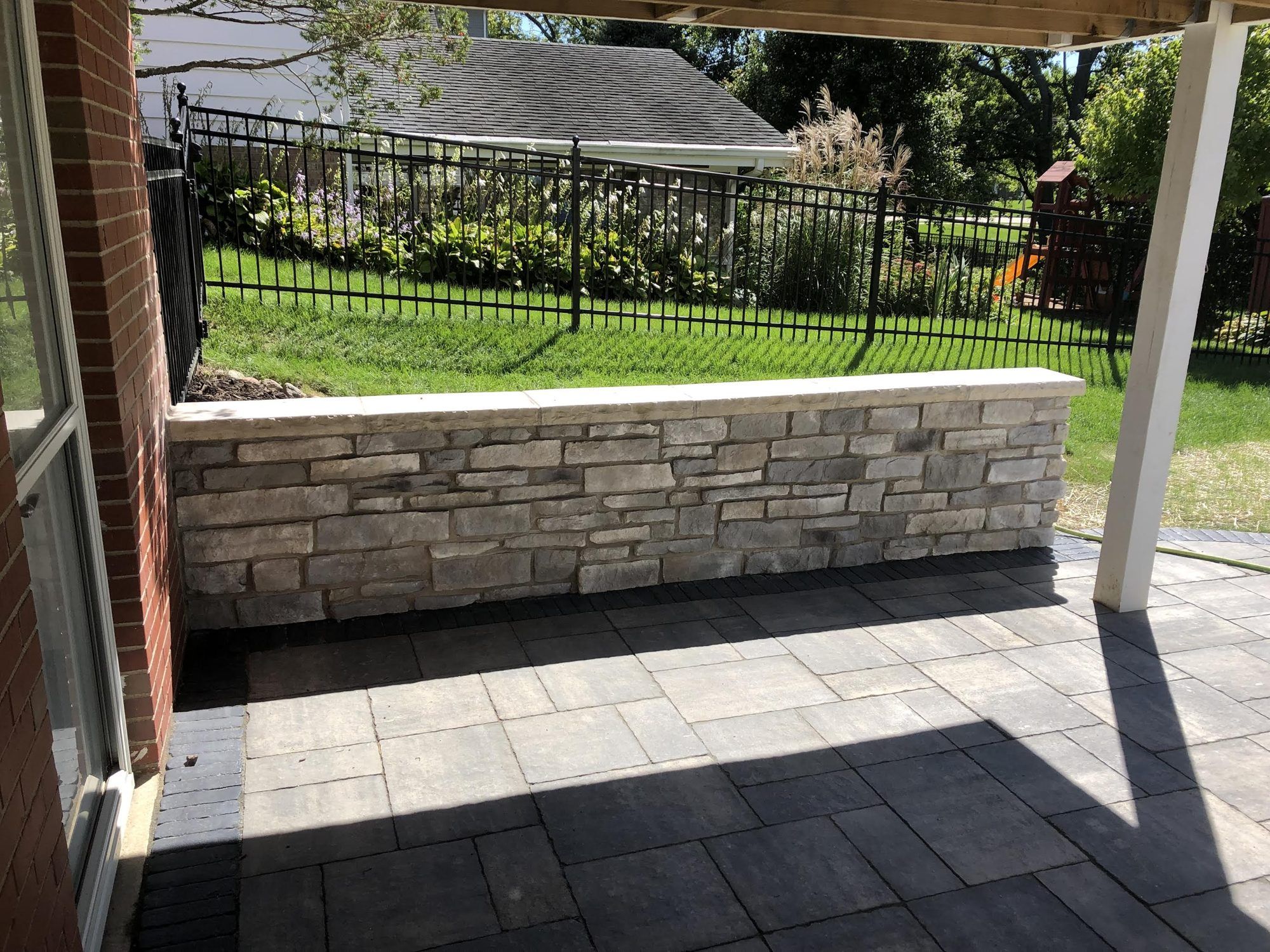 Stone seat wall and paver patio under a covered porch beside an iron fence