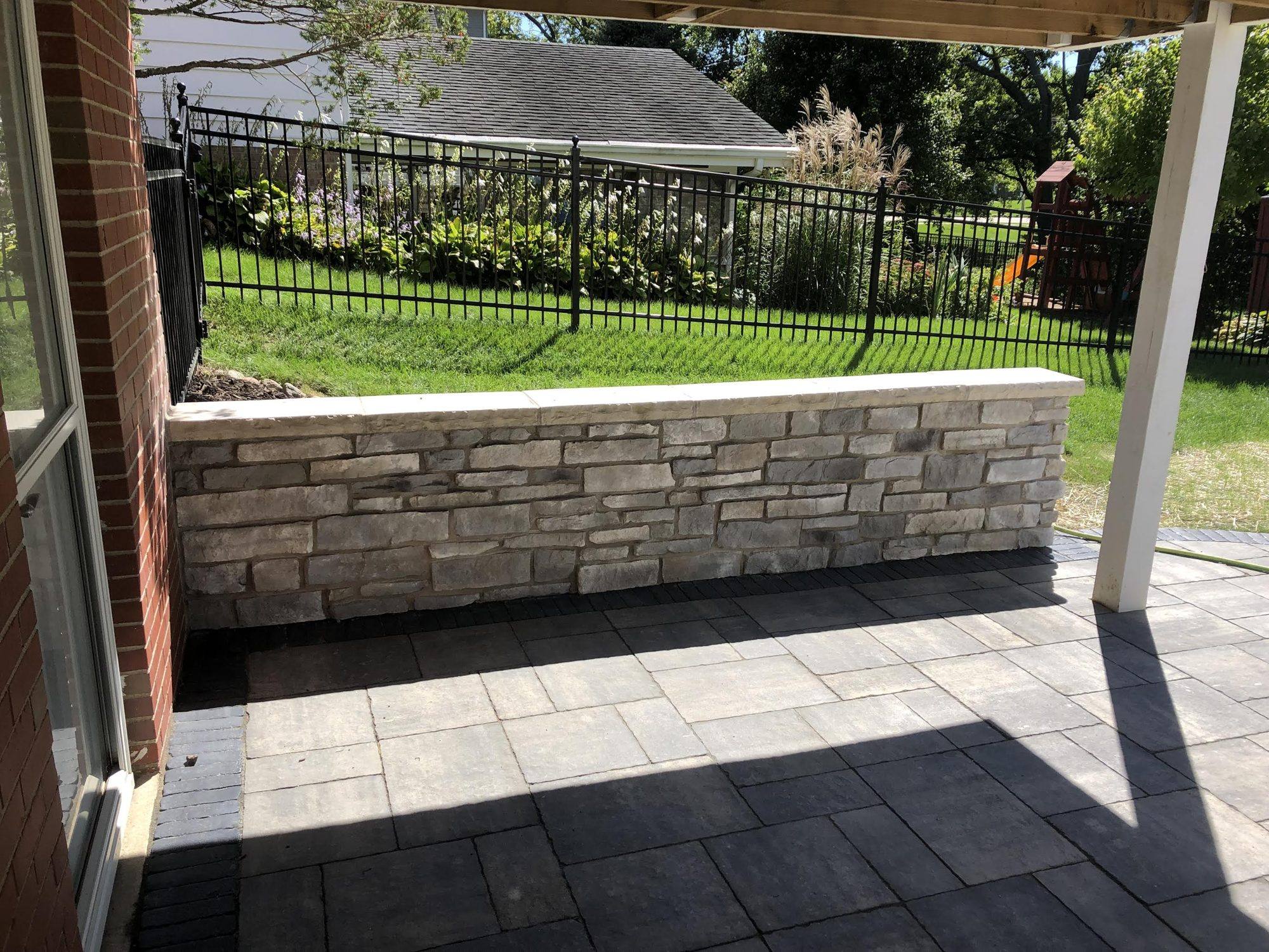 Stone seat wall and paver patio under a covered porch beside an iron fence